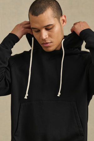 Wearing the BRODIE HOODIE by Velvet by Graham & Spencer, a person in a relaxed-fit black hoodie with white drawstrings adjusts the hood while looking down.