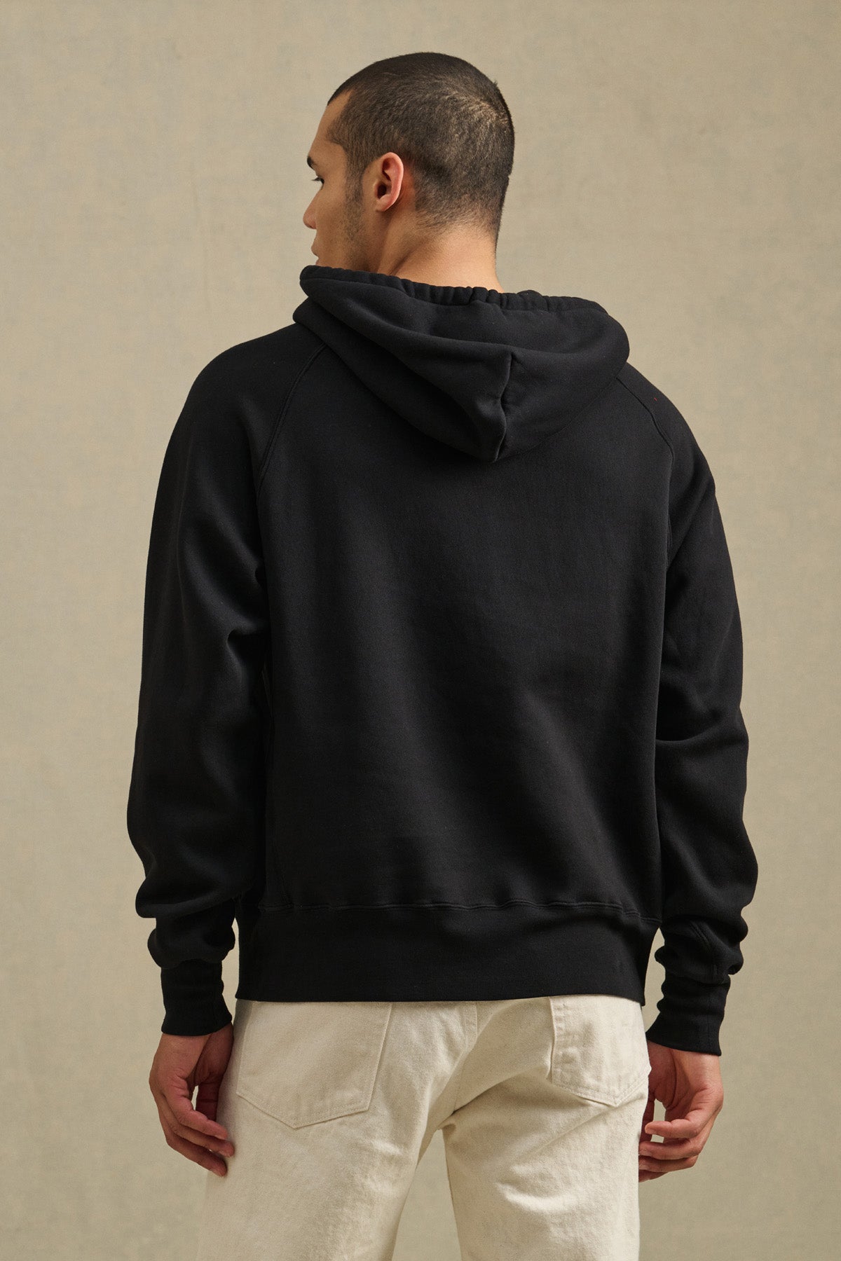 A person stands facing away, wearing the Velvet by Graham & Spencer BRODIE HOODIE in black with light-colored pants against a neutral background.
