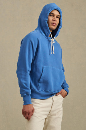 Someone models the BRODIE HOODIE by Velvet by Graham & Spencer—a blue, relaxed fit style with white drawstrings—paired with light pants against a neutral background, capturing the essence of everyday comfort.