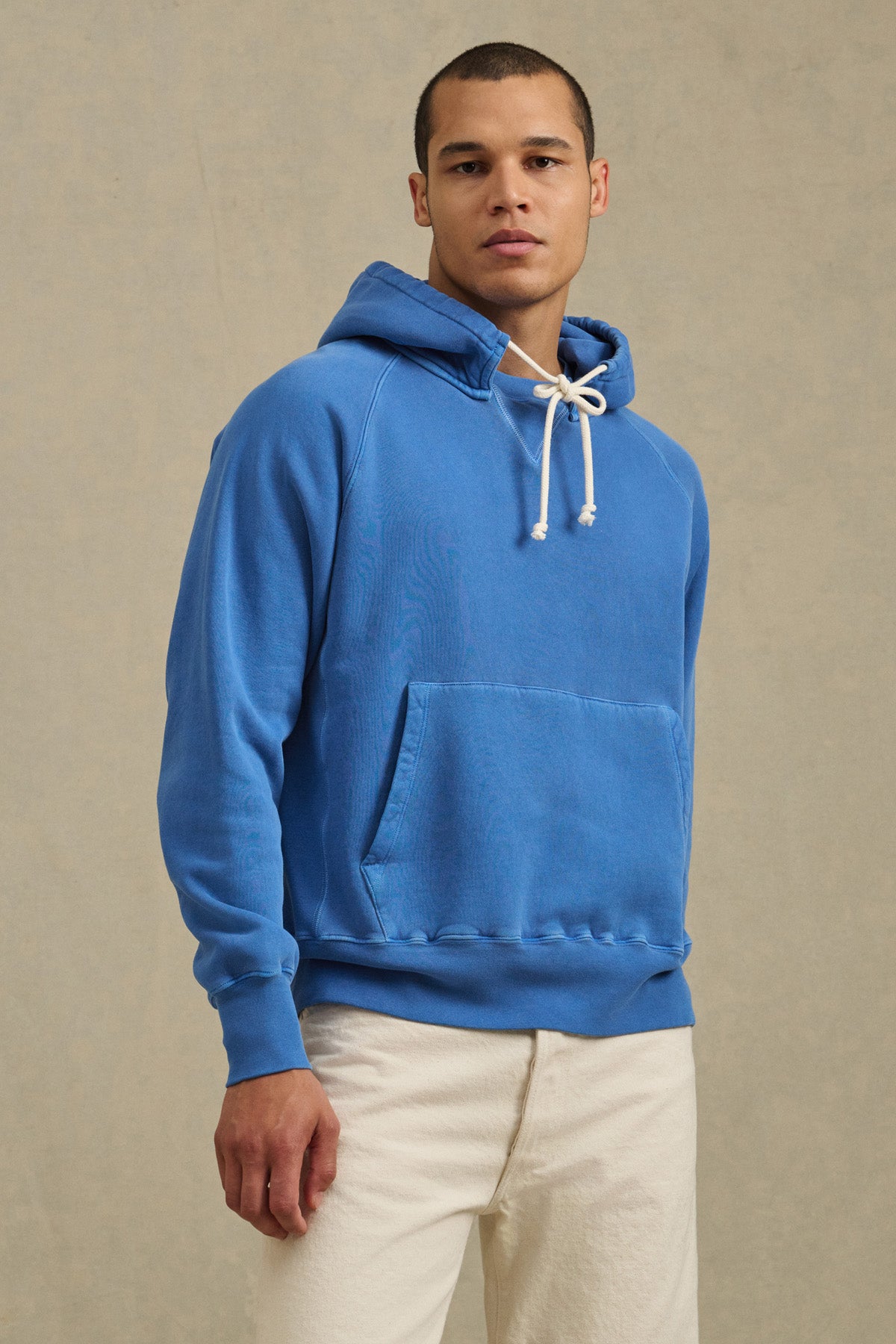 A person models the BRODIE HOODIE by Velvet by Graham & Spencer—a bright blue relaxed fit hoodie with white drawstrings—paired with light pants against a neutral background, highlighting everyday comfort.