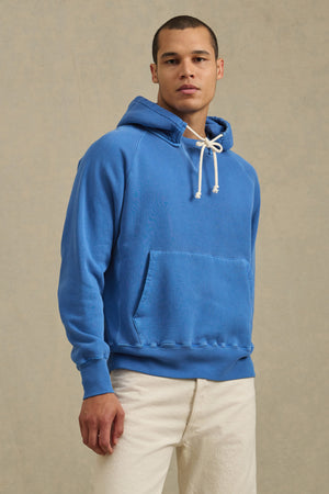 A person models the BRODIE HOODIE by Velvet by Graham & Spencer—a bright blue relaxed fit hoodie with white drawstrings—paired with light pants against a neutral background, highlighting everyday comfort.