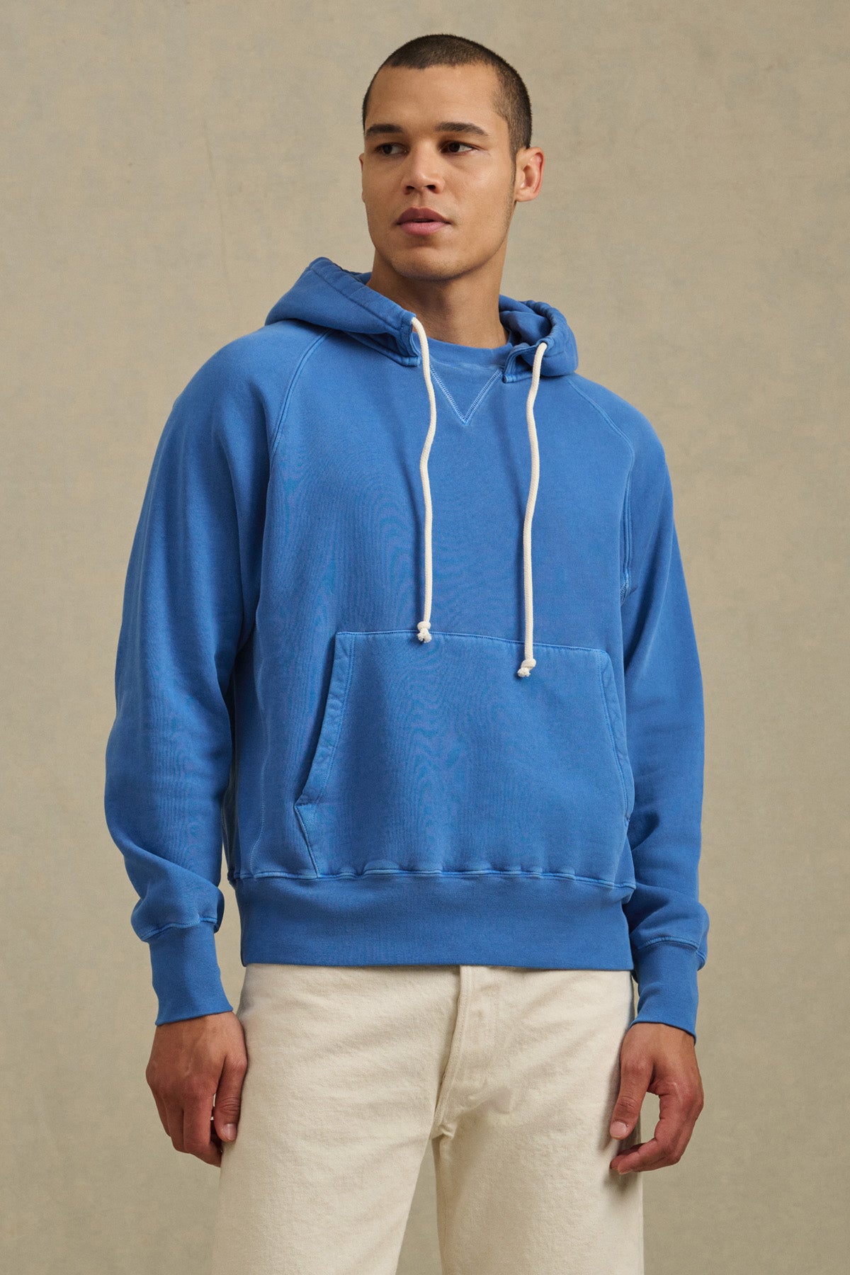 A person wearing the BRODIE HOODIE by Velvet by Graham & Spencer—a blue soft fleece hoodie with white drawstrings—and white pants stands against a neutral background, highlighting everyday comfort.