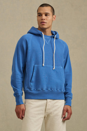 A person wearing the BRODIE HOODIE by Velvet by Graham & Spencer—a blue soft fleece hoodie with white drawstrings—and white pants stands against a neutral background, highlighting everyday comfort.