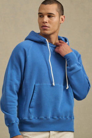 A person wears the BRODIE HOODIE by Velvet by Graham & Spencer in blue with white drawstrings and beige pants, standing against a neutral background and looking to the side, effortlessly embodying everyday comfort.