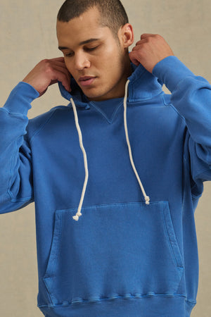 Wearing the BRODIE HOODIE by Velvet by Graham & Spencer in blue with white drawstrings, a person adjusts the hood, enjoying soft fleece and everyday comfort against a plain backdrop.