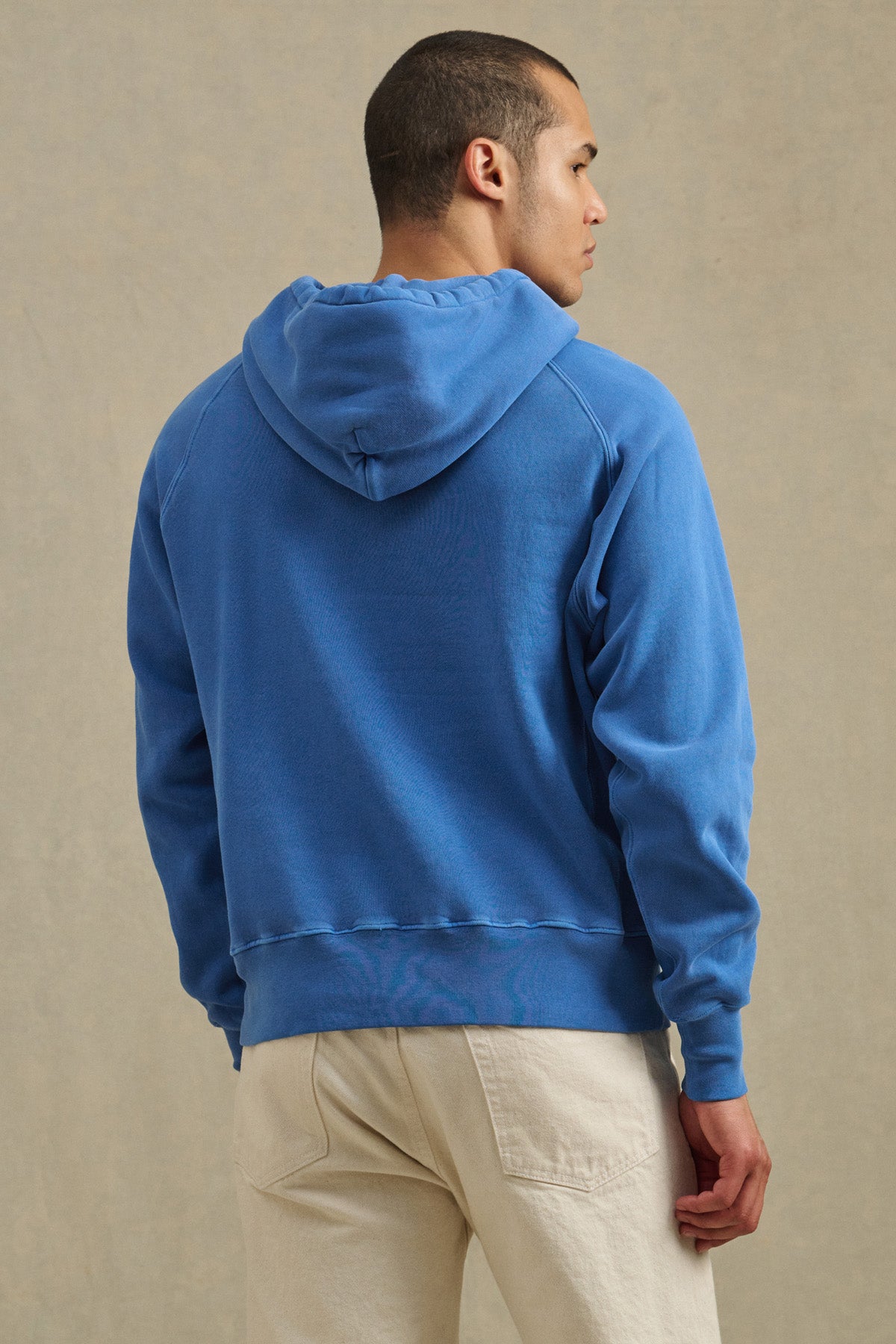 A person wearing the Velvet by Graham & Spencer BRODIE HOODIE in blue and beige pants stands facing away from the camera against a light background, highlighting this soft fleece for everyday comfort.-41145219580097