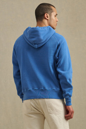 A person wearing the Velvet by Graham & Spencer BRODIE HOODIE in blue and beige pants stands facing away from the camera against a light background, highlighting this soft fleece for everyday comfort.