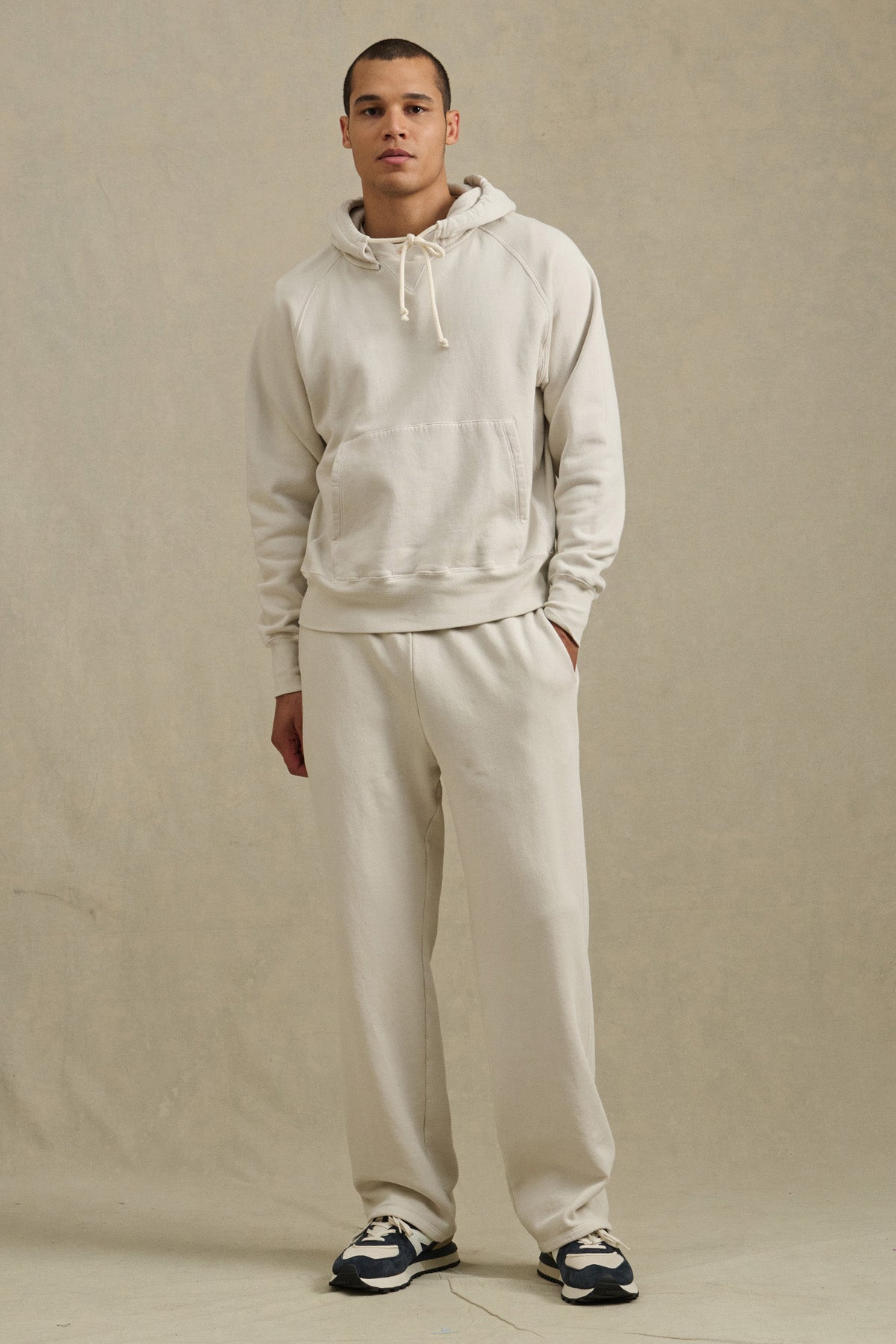 A person stands against a neutral background wearing the BRODIE HOODIE in light beige from Velvet by Graham & Spencer, paired with matching sweatpants and black-and-white sneakers.-40850658066625