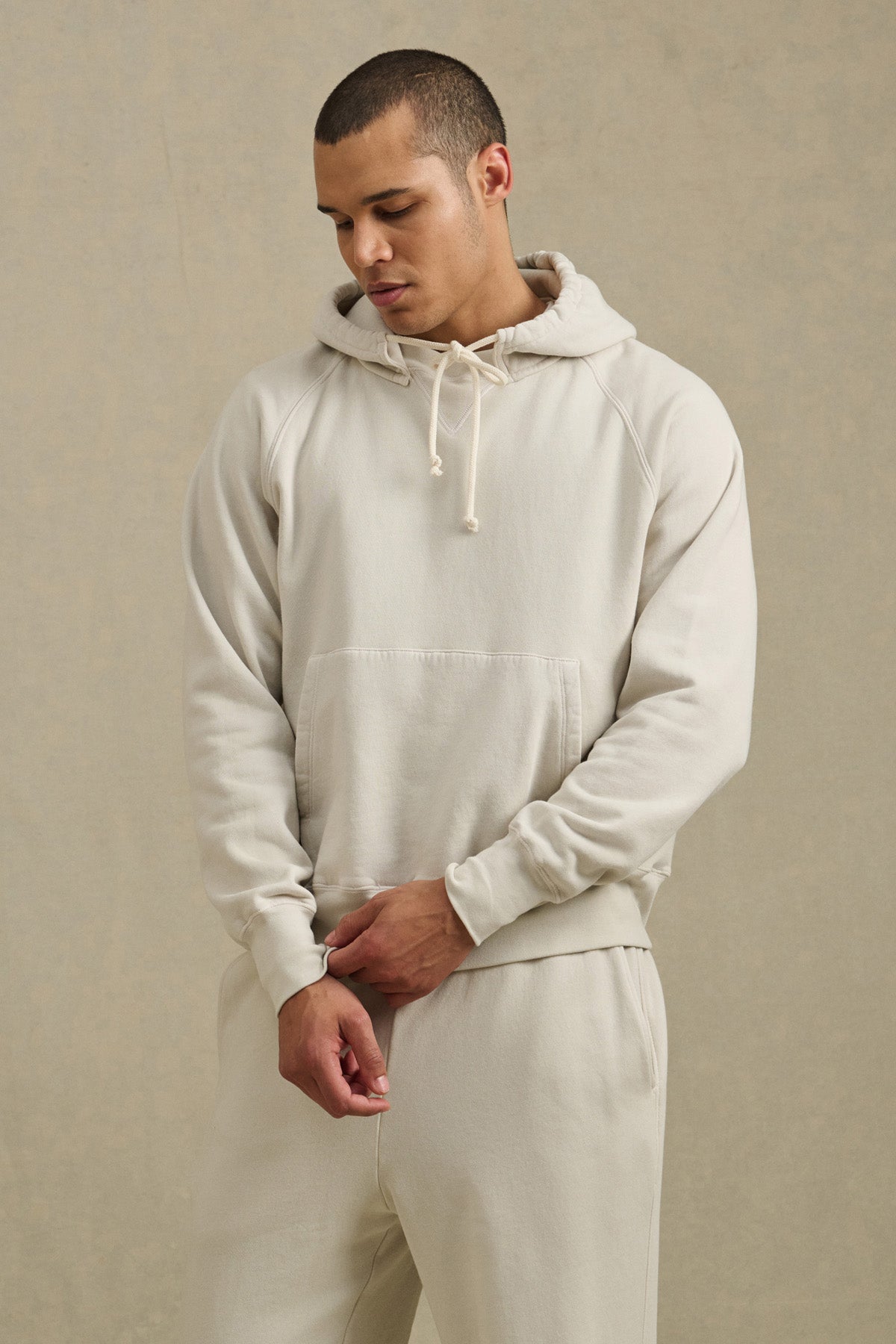 A man wearing the BRODIE HOODIE by Velvet by Graham & Spencer in light beige with matching sweatpants stands against a neutral background, looking downward with his hands in front of him.