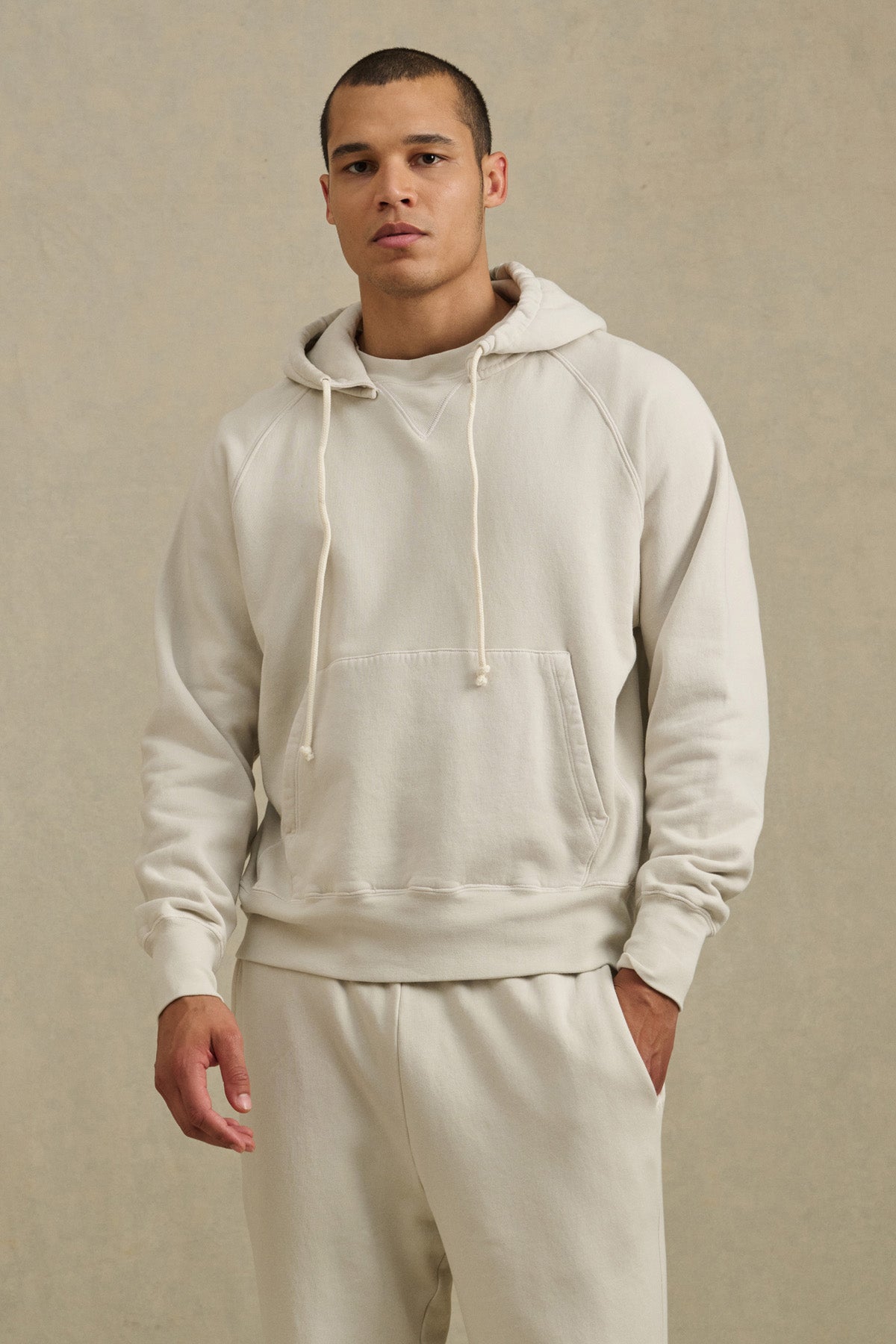 A person wears the BRODIE HOODIE by Velvet by Graham & Spencer—a relaxed fit, light beige hoodie—paired with matching sweatpants, standing against a neutral background and looking at the camera with one hand in their pocket.