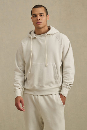 A person wears the BRODIE HOODIE by Velvet by Graham & Spencer—a relaxed fit, light beige hoodie—paired with matching sweatpants, standing against a neutral background and looking at the camera with one hand in their pocket.