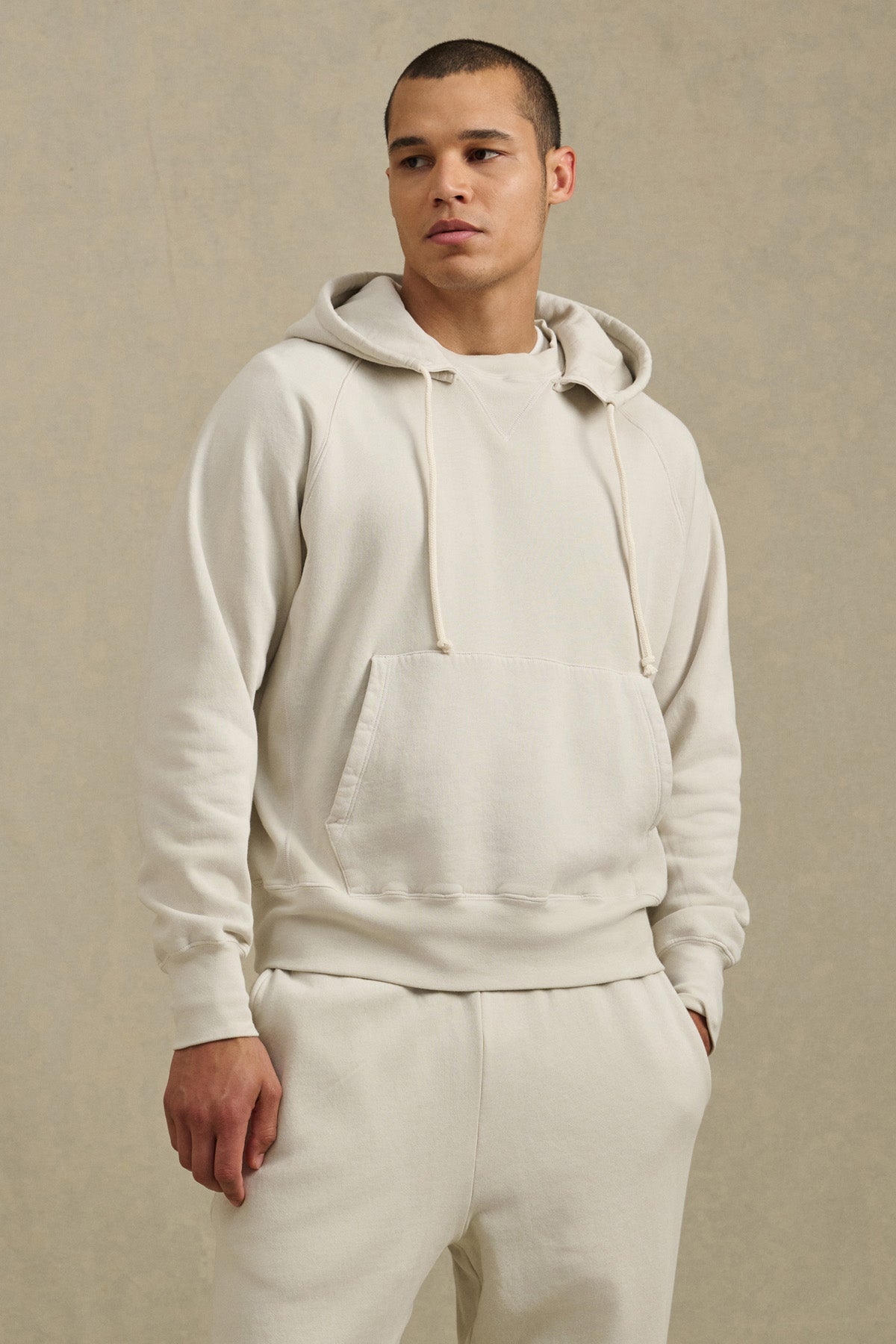A person wears the BRODIE HOODIE by Velvet by Graham & Spencer—crafted in soft cream fleece—paired with matching sweatpants, standing against a neutral background and looking to the side.-40850657870017