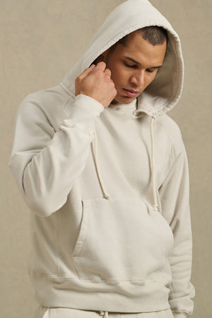 A man wears the BRODIE HOODIE by Velvet by Graham & Spencer, a light beige, relaxed-fit fleece hoodie with drawstrings, as he looks down and touches the edge of his hood.