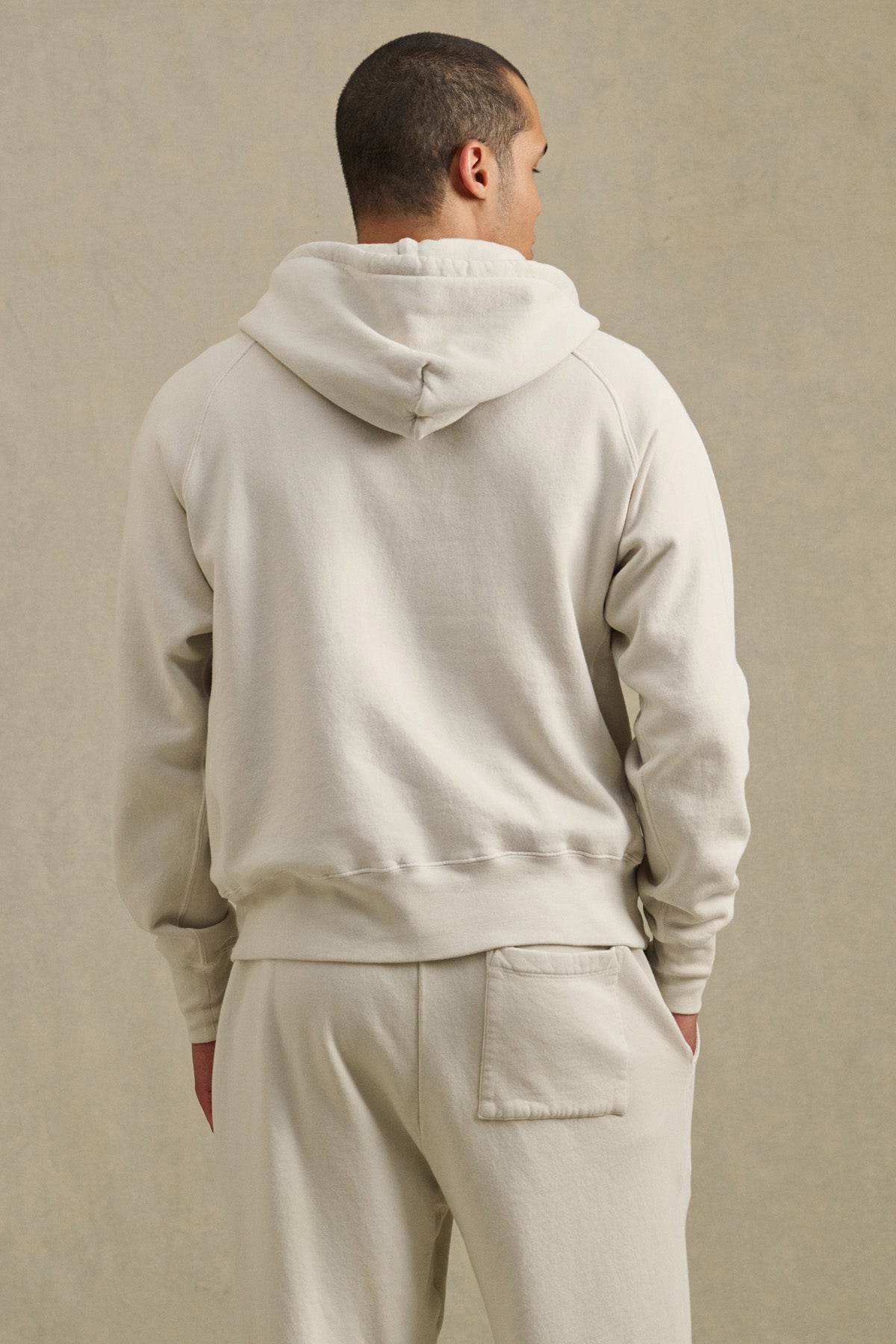 A person stands with their back to the camera against a neutral background, wearing the cream-colored BRODIE HOODIE by Velvet by Graham & Spencer and matching soft fleece sweatpants in a relaxed fit.-40850657706177