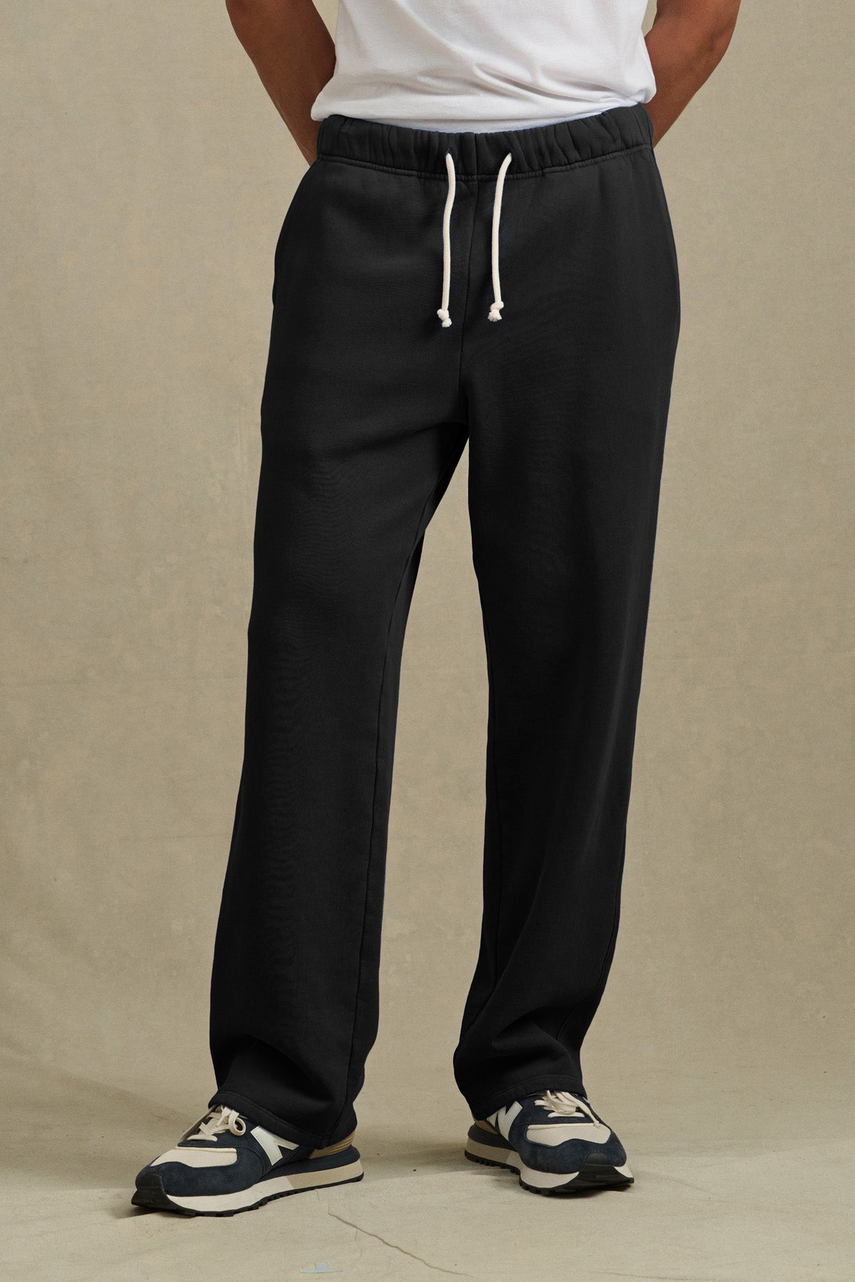 A person wears Velvet by Graham & Spencer’s HARLAND SWEATPANT in black, featuring a drawstring elastic waist, paired with a white t-shirt and black-and-white sneakers, standing against a plain beige background.-40850732613825