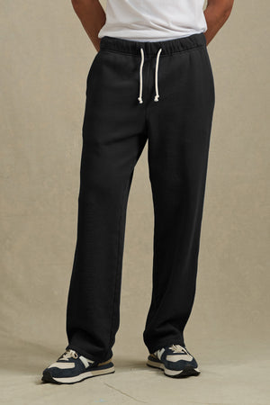 A person wears Velvet by Graham & Spencer’s HARLAND SWEATPANT in black, featuring a drawstring elastic waist, paired with a white t-shirt and black-and-white sneakers, standing against a plain beige background.