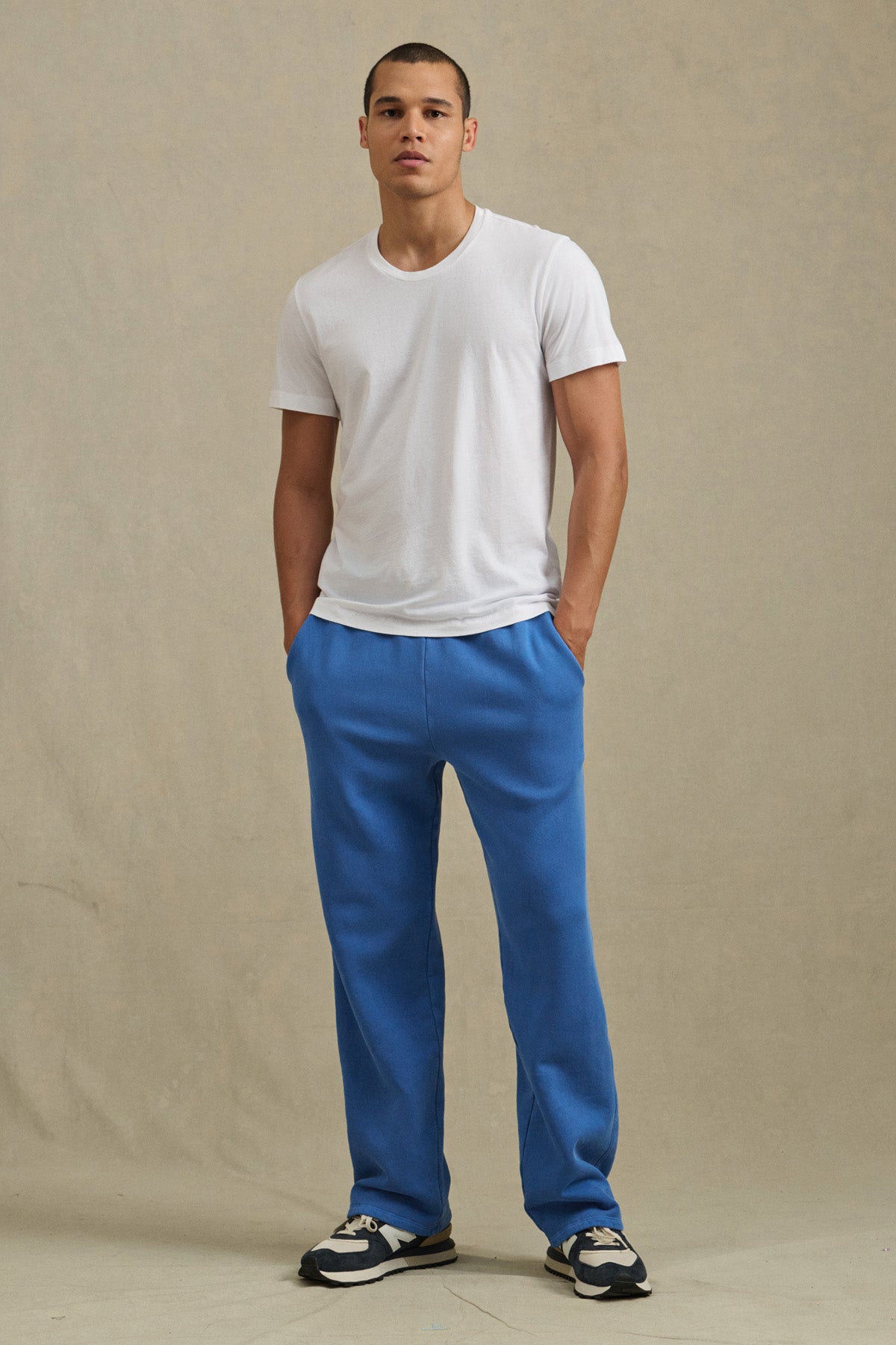 A person stands against a plain background wearing a white T-shirt, black-and-white sneakers, and blue HARLAND SWEATPANT by Velvet by Graham & Spencer, featuring straight legs and a drawstring elastic waist, with hands in pockets.-41145350652097
