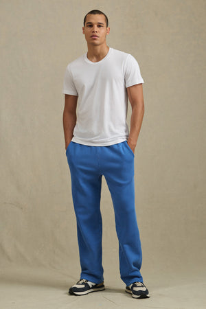 A person stands against a plain background wearing a white T-shirt, black-and-white sneakers, and blue HARLAND SWEATPANT by Velvet by Graham & Spencer, featuring straight legs and a drawstring elastic waist, with hands in pockets.