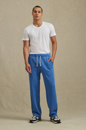 A person stands facing forward wearing Velvet by Graham & Spencer's HARLAND SWEATPANT in blue, featuring a drawstring elastic waist, paired with a white t-shirt and black-and-white sneakers against a plain beige background.