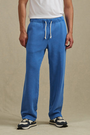 A person wears a white T-shirt, Velvet by Graham & Spencer HARLAND SWEATPANT in blue with a drawstring waist, and black-and-white sneakers, standing against a beige background.