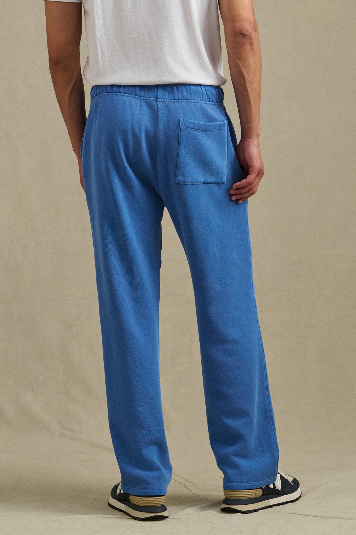 A man stands on a beige surface, shown from the back, wearing a white T-shirt, HARLAND SWEATPANT by Velvet by Graham & Spencer—blue straight-leg pants with a back pocket and drawstring elastic waist—and black and white sneakers.