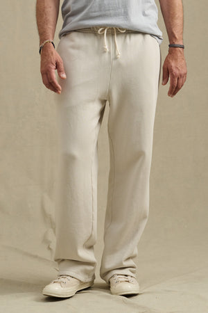 A person wears the HARLAND SWEATPANT by Velvet by Graham & Spencer in off-white, paired with a light grey t-shirt and white sneakers, standing against a beige background.