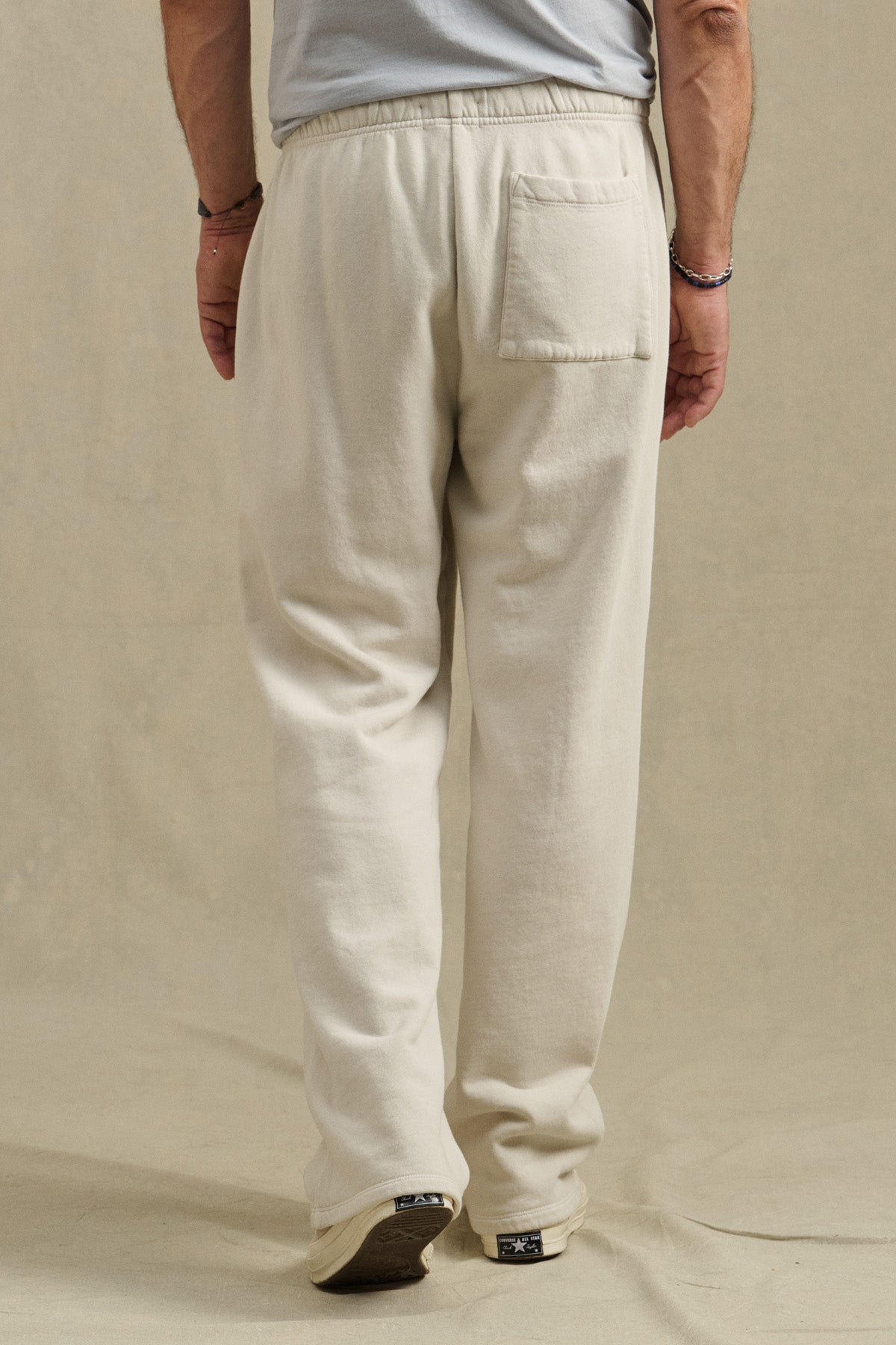 A person stands on a beige surface wearing Velvet by Graham & Spencer’s HARLAND SWEATPANT in off-white, featuring a drawstring waist, single back pocket, midweight fleece fabric, and relaxed fit, paired with a gray shirt.-40850732449985