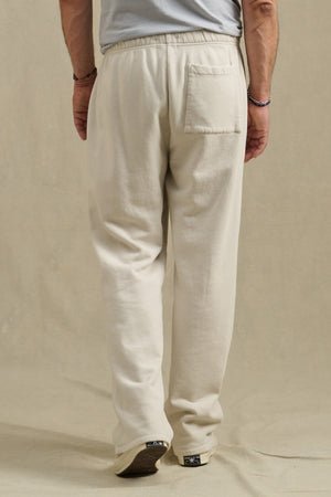A person stands on a beige surface wearing Velvet by Graham & Spencer’s HARLAND SWEATPANT in off-white, featuring a drawstring waist, single back pocket, midweight fleece fabric, and relaxed fit, paired with a gray shirt.