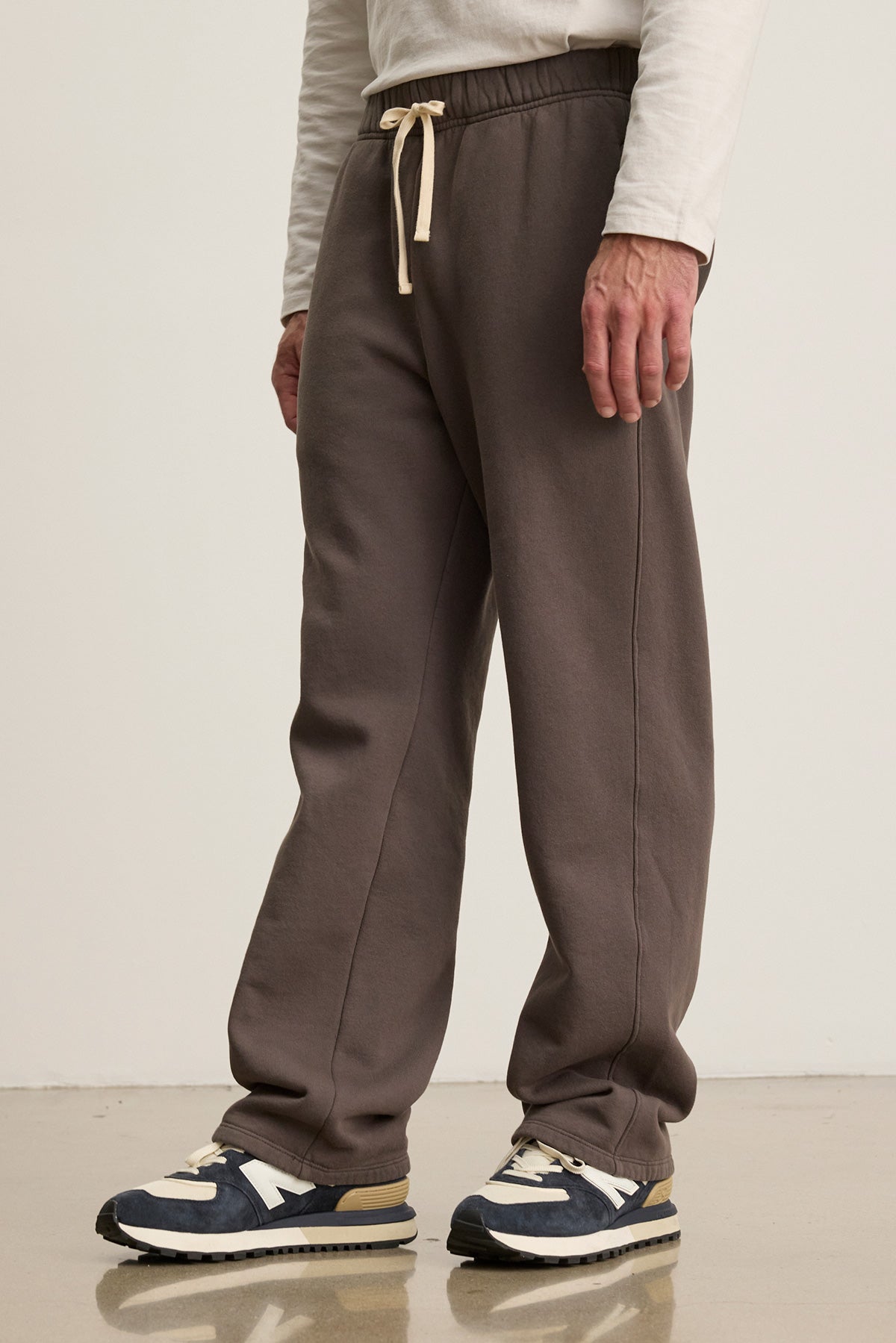 A person wears the HUDSON SWEATPANT in brown by Velvet by Graham & Spencer, paired with a white long-sleeve shirt and black-and-white sneakers on a polished floor—ideal for comfortable everyday wear.-40276964638913
