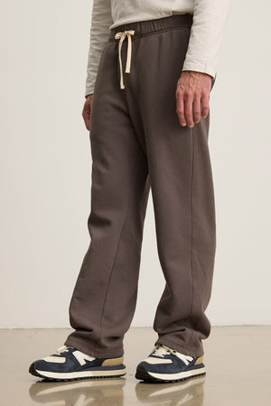A person wears the HUDSON SWEATPANT in brown by Velvet by Graham & Spencer, paired with a white long-sleeve shirt and black-and-white sneakers on a polished floor—ideal for comfortable everyday wear.