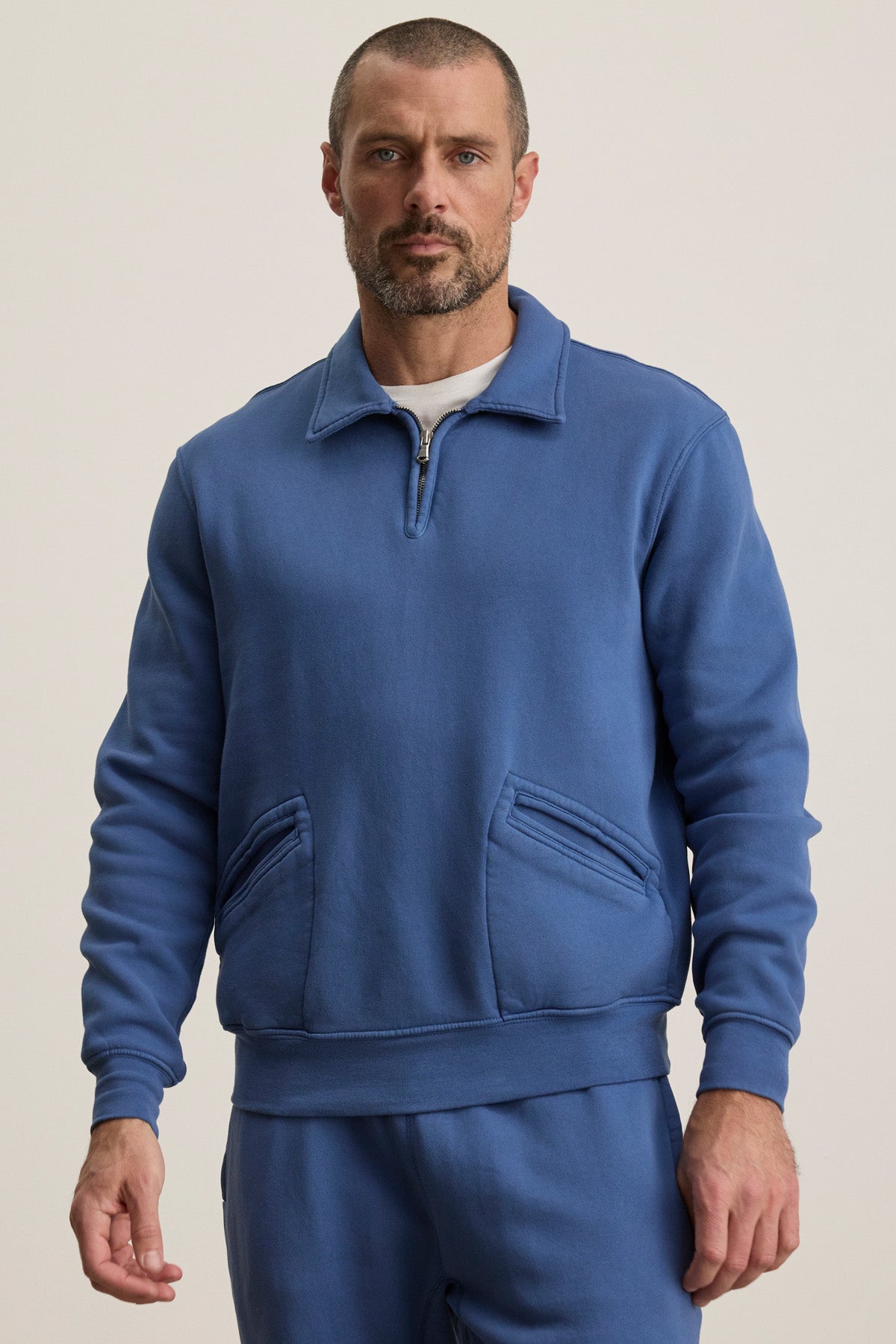 A man wears the LEVI SWEATSHIRT by Velvet by Graham & Spencer, a blue quarter zip with front pockets, paired with matching blue pants, standing against a plain light background.-40606036459713