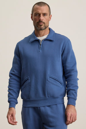 A man wears the LEVI SWEATSHIRT by Velvet by Graham & Spencer, a blue quarter zip with front pockets, paired with matching blue pants, standing against a plain light background.