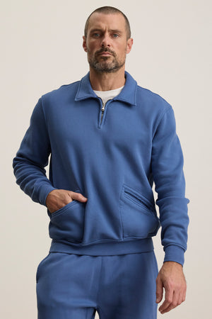 A man stands against a plain light background with one hand in his pocket, wearing the blue LEVI SWEATSHIRT and matching sweatpants by Velvet by Graham & Spencer.