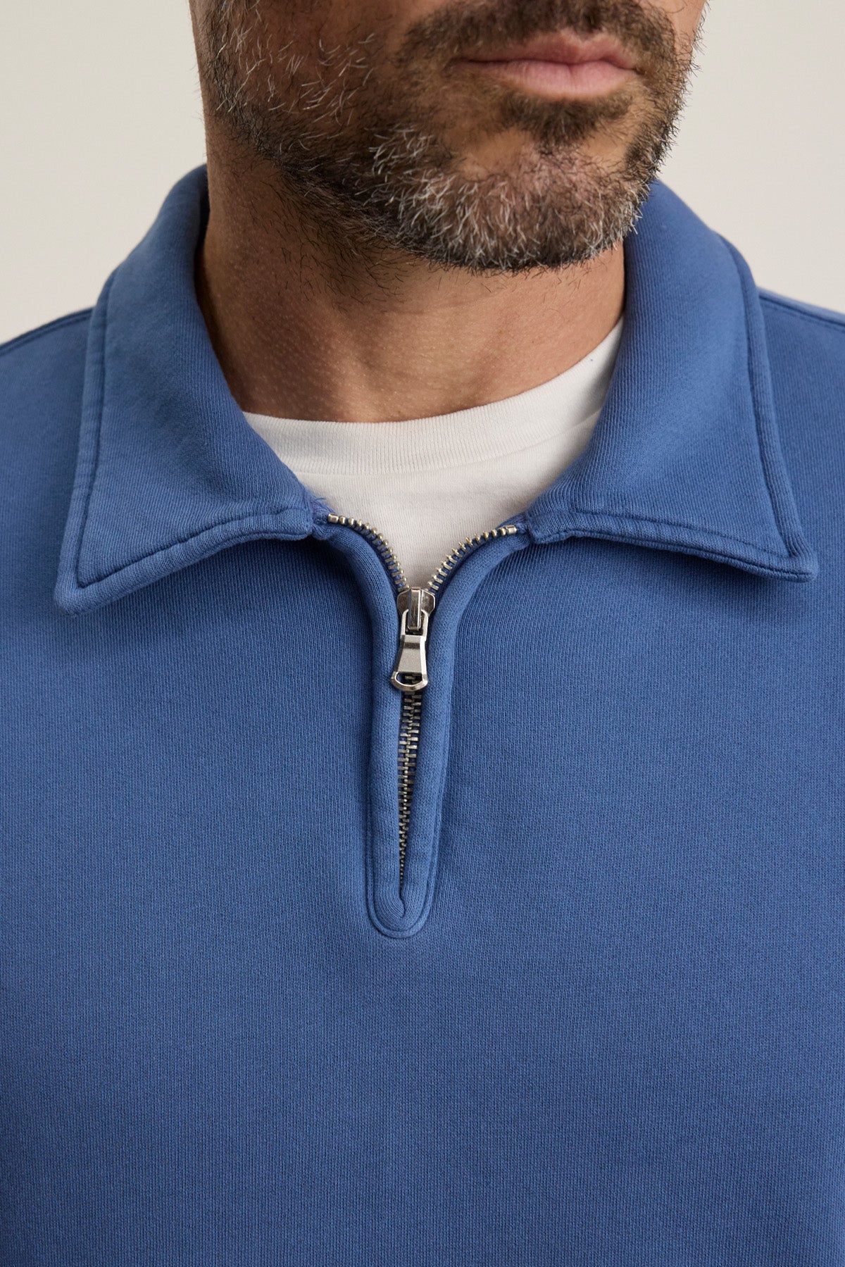 A close-up shows a man with facial hair wearing the LEVI SWEATSHIRT by Velvet by Graham & Spencer—a vintage-inspired blue quarter zip styled over a white t-shirt, highlighting the zipper and part of his face.-40606036295873