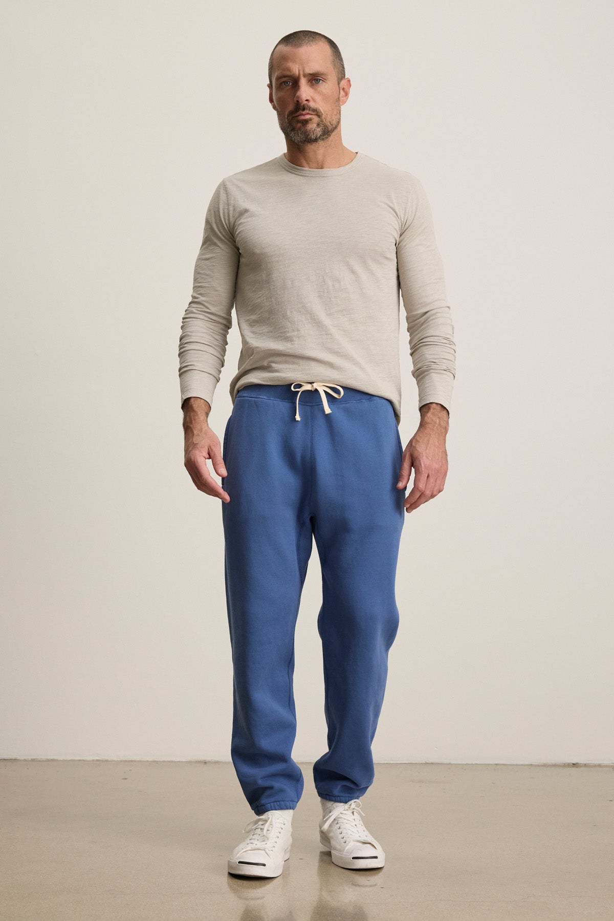A man stands indoors against a plain background wearing a light beige long-sleeve shirt, Velvet by Graham & Spencer NORRIS JOGGER heavyweight blue fleece sweatpants with a drawstring elastic waist, and white sneakers.-40606036754625