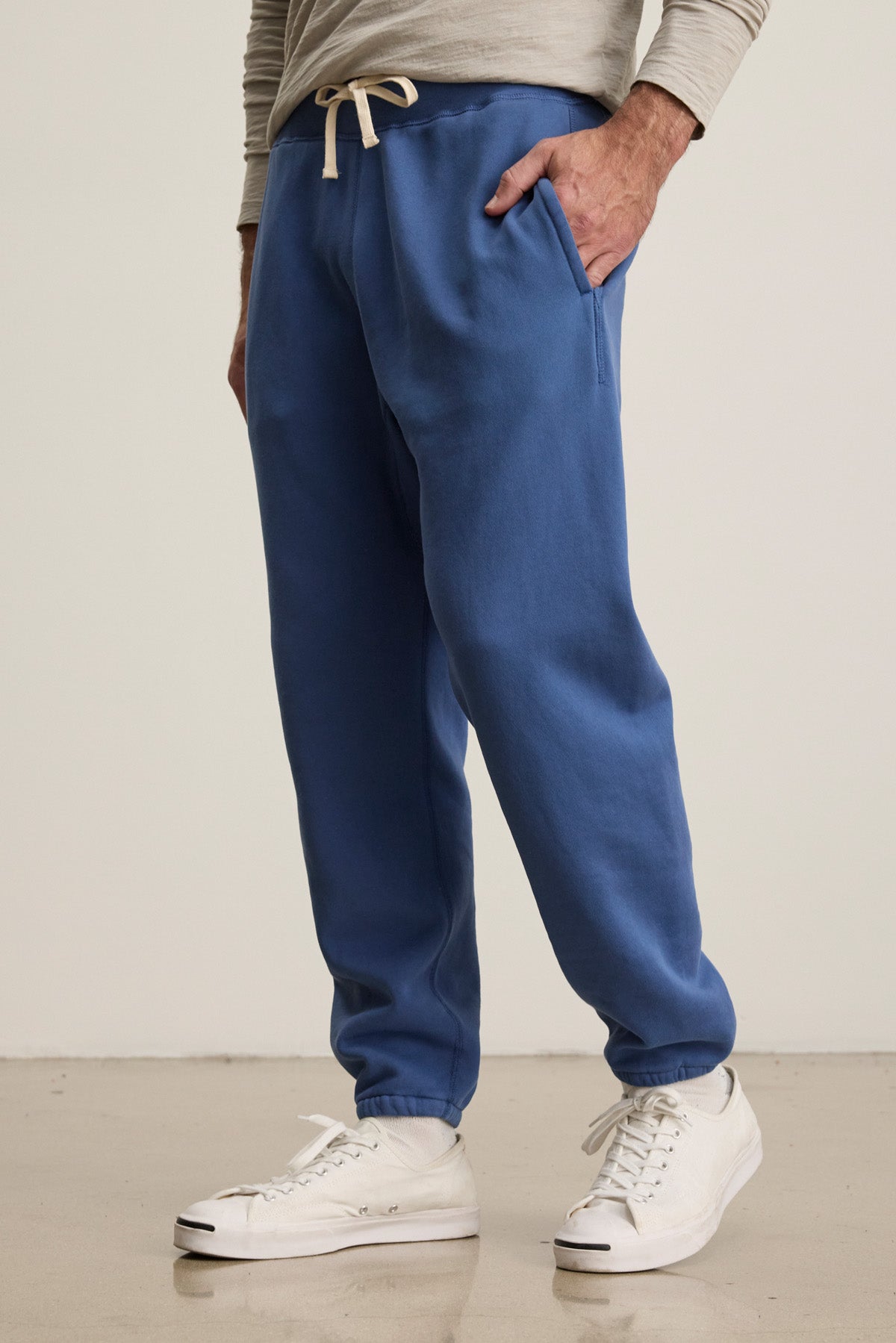Wearing Velvet by Graham & Spencer's NORRIS JOGGER, a person stands with hands in pockets. The blue fleece jogger features a drawstring elastic waist, pairing perfectly with a light long-sleeve shirt and white high-top sneakers for everyday style.-40606036689089