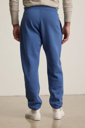 A person faces away, wearing Velvet by Graham & Spencer's NORRIS JOGGER—blue heavyweight fleece sweatpants with a drawstring waist—paired with a beige long-sleeve shirt and white sneakers on a plain light-colored floor.