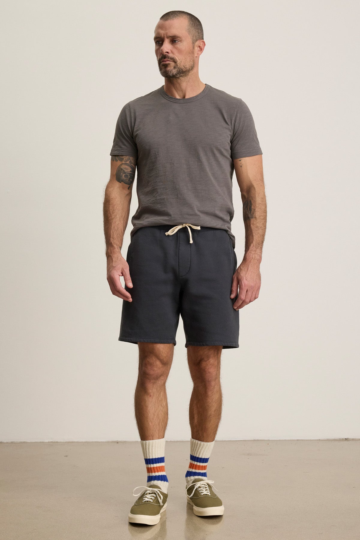 A man stands indoors against a plain light-colored wall wearing a gray t-shirt, RUDY SHORT by Velvet by Graham & Spencer in navy, with a drawstring waist, paired with striped socks and olive green sneakers.
