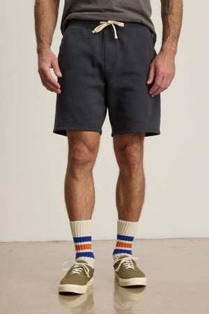 A person wearing a gray T-shirt, Velvet by Graham & Spencer RUDY SHORTS with a drawstring waist, striped socks, and olive green sneakers stands on a smooth floor.