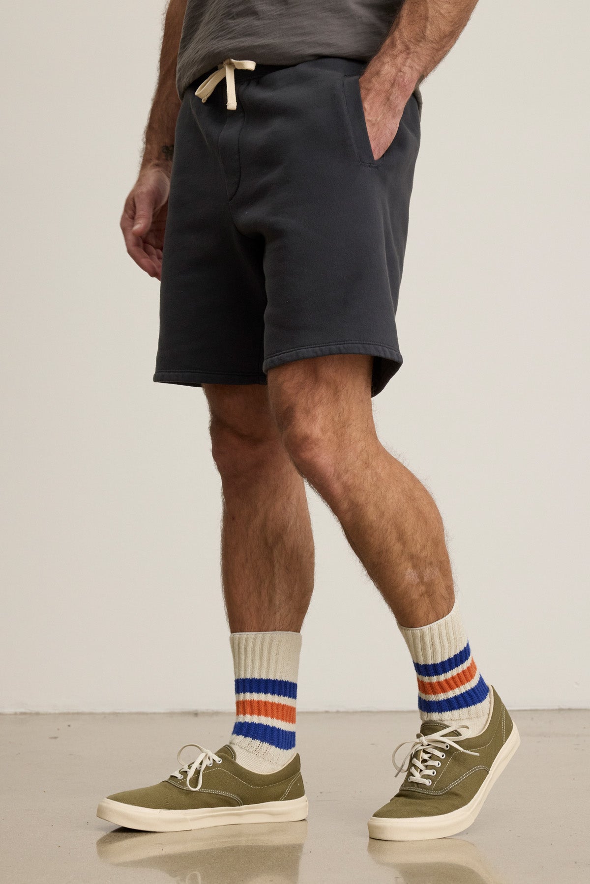 Wearing Velvet by Graham & Spencer RUDY SHORTS, a grey shirt, olive green sneakers, and cream socks with orange and blue stripes, a person stands on a light floor.-40606042816705