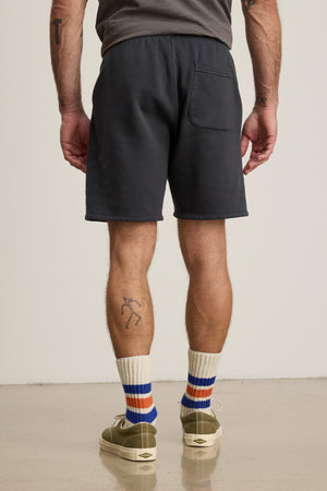 A person faces away, wearing a gray tee and RUDY SHORT lounge shorts by Velvet by Graham & Spencer, with a drawstring waist, striped socks, green shoes, and a stick-figure tattoo on their left calf.