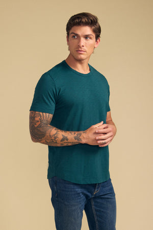 A man with a tattooed right arm wears the AMARO TEE by Velvet by Graham & Spencer—a green Peruvian cotton slub crewneck—paired with blue jeans, standing against a beige background with his hands clasped; ideal for layering.