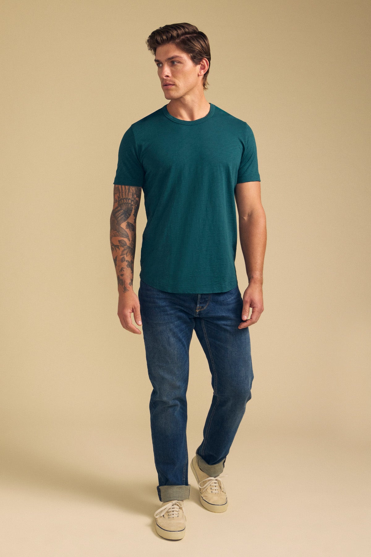 A man stands against a beige background wearing the AMARO TEE by Velvet by Graham & Spencer—a green Peruvian cotton slub crewneck—paired with blue jeans and beige sneakers. A tattoo is visible on one arm as he looks slightly to his left.-41701717409985