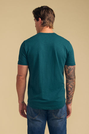 A man faces away from the camera in a teal AMARO TEE by Velvet by Graham & Spencer, made from Peruvian cotton slub with a crew neckline, paired with blue jeans; a large tattoo covers his right arm against a beige background.