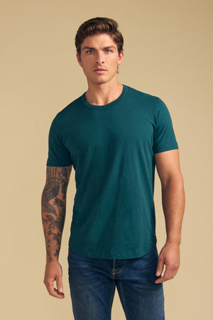 A man with light brown hair and a tattoo sleeve wears the Velvet by Graham & Spencer AMARO TEE in teal Peruvian cotton slub, featuring a classic crew neckline, styled with blue jeans against a beige background. Ideal as a layering piece.