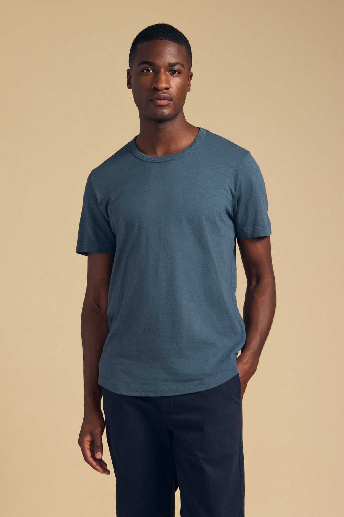 A man wearing the AMARO TEE in slate blue and dark trousers.