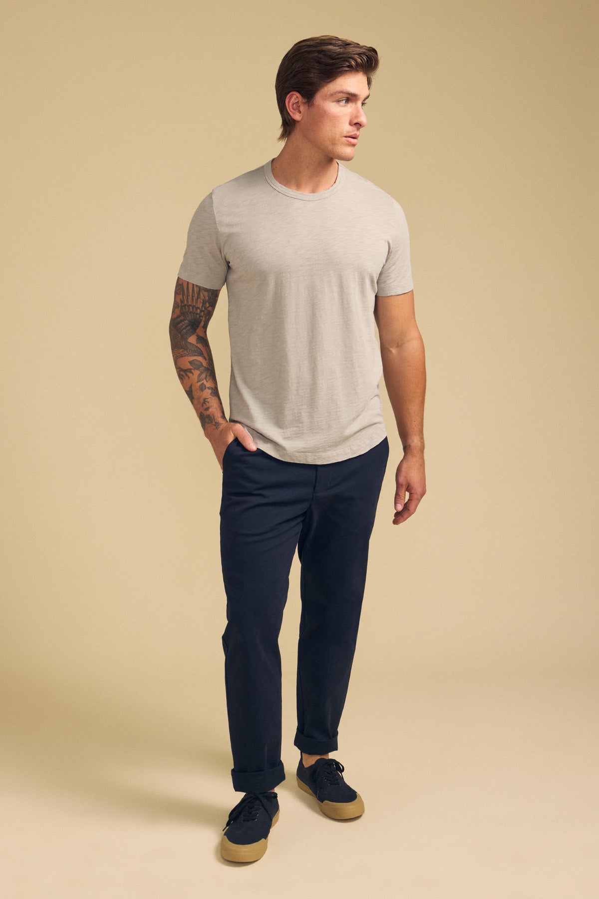 A man with a tattoo on his right arm wears the AMARO TEE by Velvet by Graham & Spencer—a light grey Peruvian cotton slub crewneck—paired with dark pants and black sneakers, standing against a plain beige background.-41701717049537