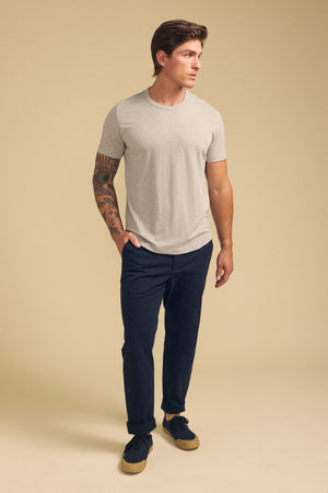 A man with a tattoo on his right arm wears the AMARO TEE by Velvet by Graham & Spencer—a light grey Peruvian cotton slub crewneck—paired with dark pants and black sneakers, standing against a plain beige background.