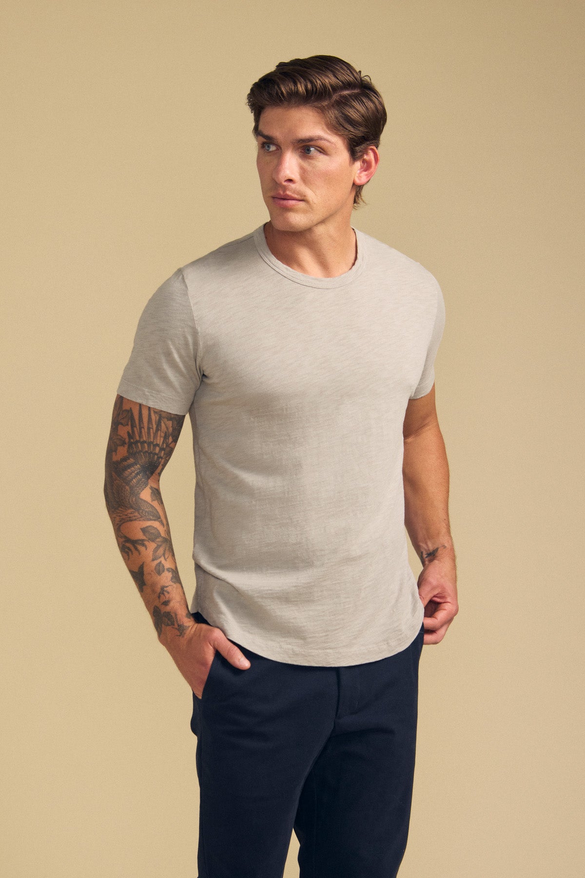 Man wearing the AMARO TEE by Velvet by Graham & Spencer—a light beige Peruvian cotton slub short-sleeve t-shirt with a classic crew neck—paired with dark pants, hands in pockets, looking to the right against a neutral background.