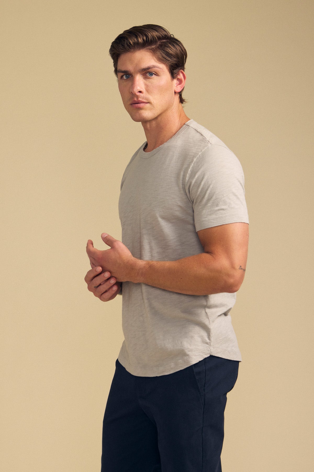 A man with short brown hair wears the AMARO TEE by Velvet by Graham & Spencer—a light grey Peruvian cotton slub crewneck—paired with dark pants, standing against a beige background and looking at the camera.-41701716852929